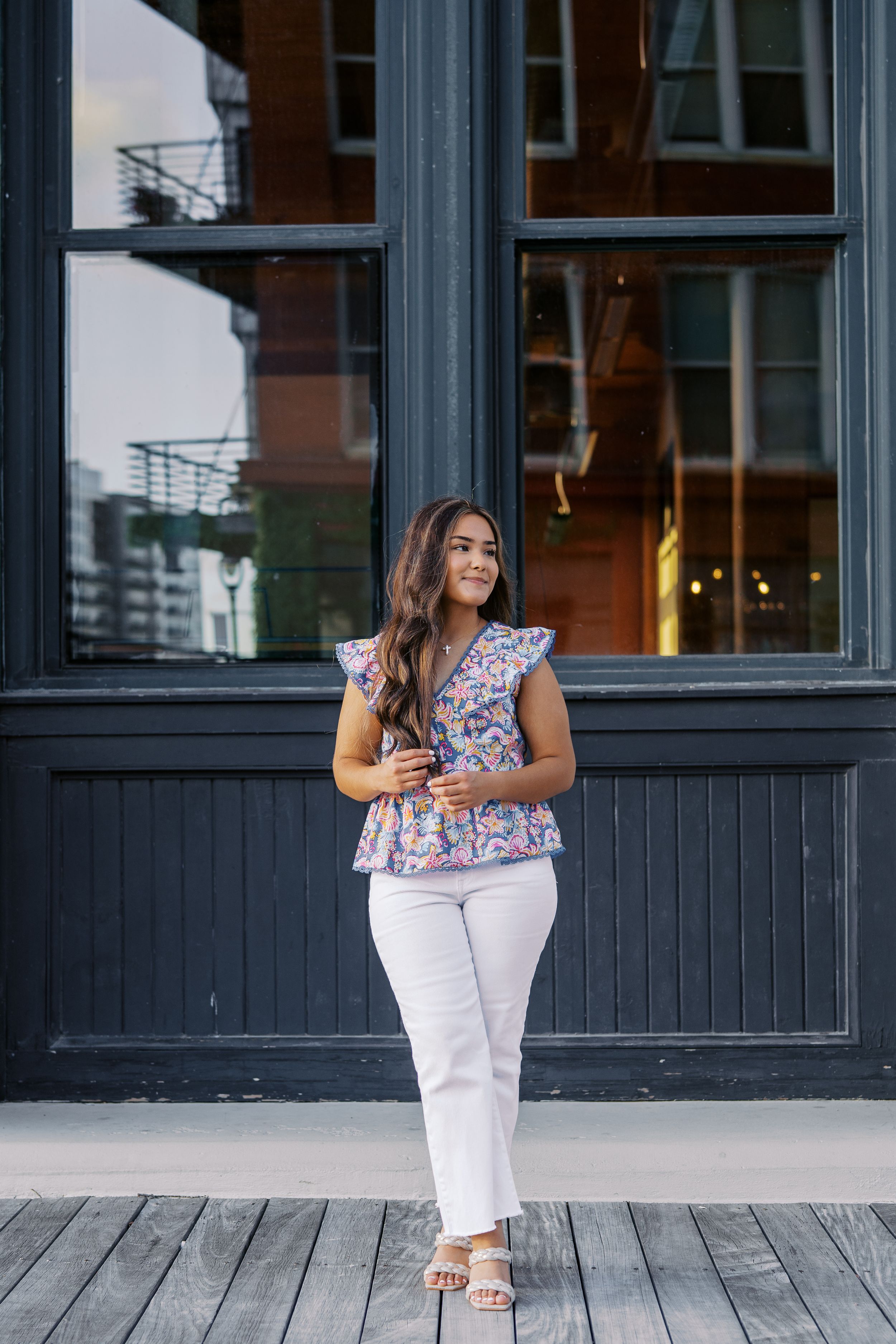 Standing by dark storefront windows wearing white pants and floral patterned top.