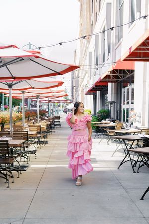 A flowing pink dress captured in motion along a sidewalk with outdoor cafe seating and umbrellas.