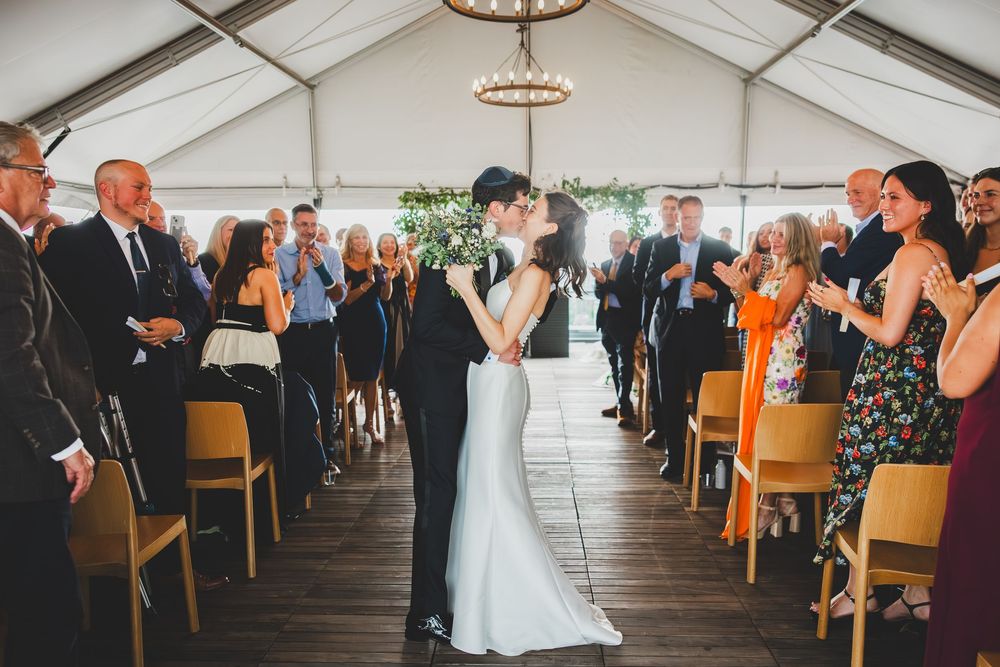 Wedding guests stand and applaud as the newlyweds walk down the aisle at an indoor ceremony.