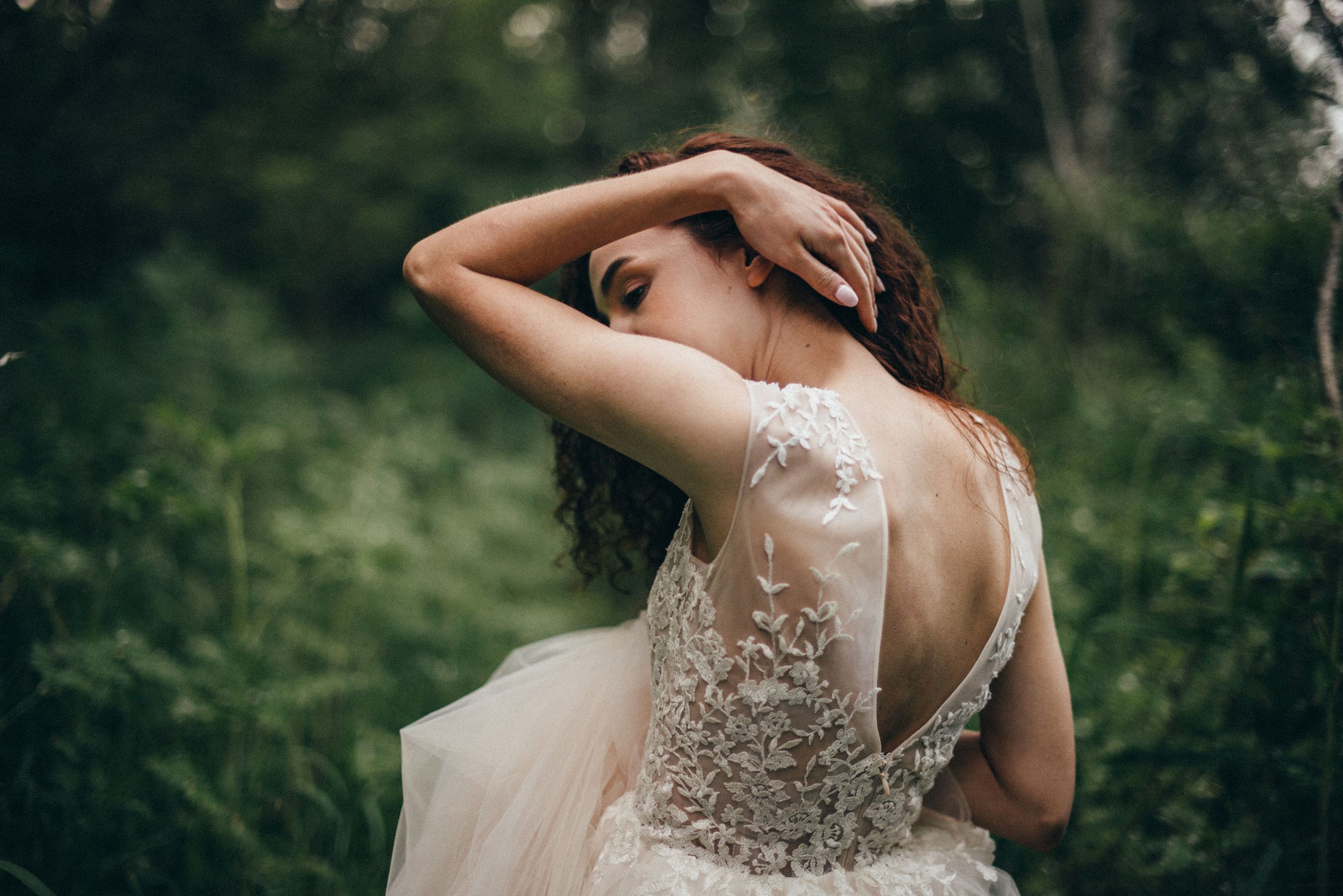 A bride in a lace backless wedding gown poses gracefully among lush green foliage.