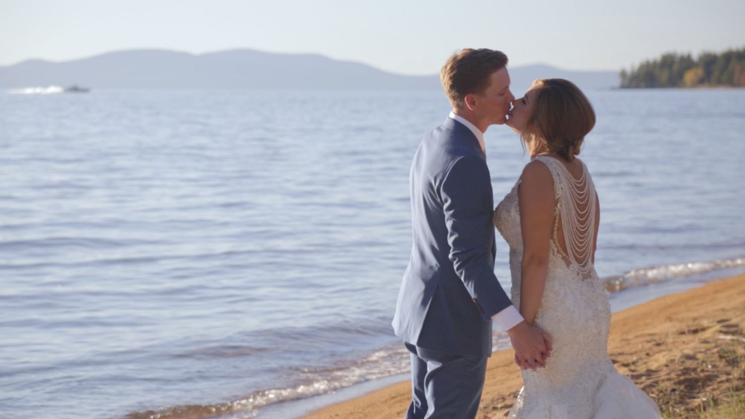 A couple shares a romantic kiss on a sandy lakeside beach, the bride in a lace gown and groom in a gray suit.