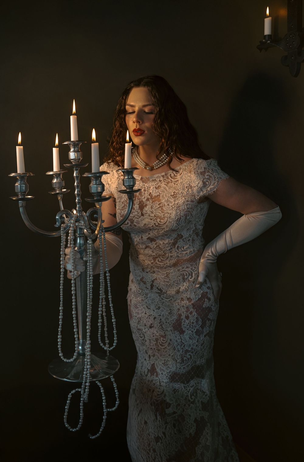 Elegant figure in lace dress poses with vintage candelabra in dark atmospheric setting.