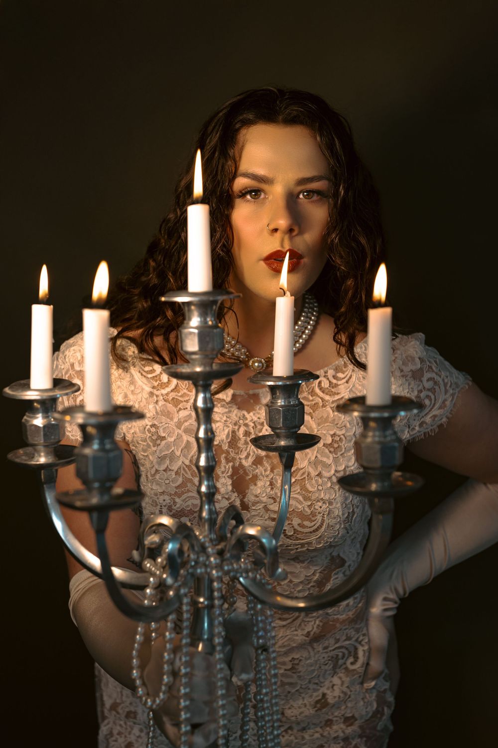 Person in vintage lace dress holds ornate candelabra with lit candles in moody dark setting.