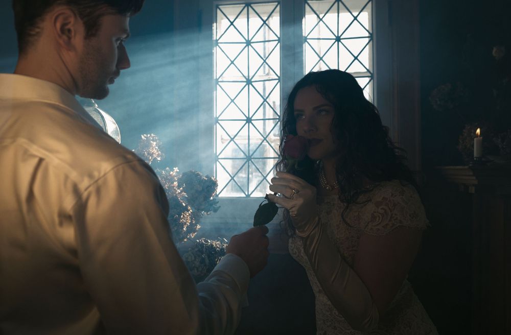 Two figures stand in a dimly lit room with cross-patterned windows casting atmospheric light and shadows.