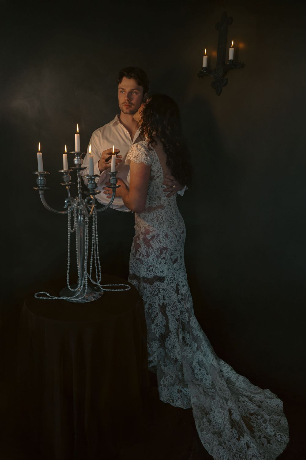 A figure in a flowing lace gown poses dramatically near elegant candelabras in a dark, moody setting.