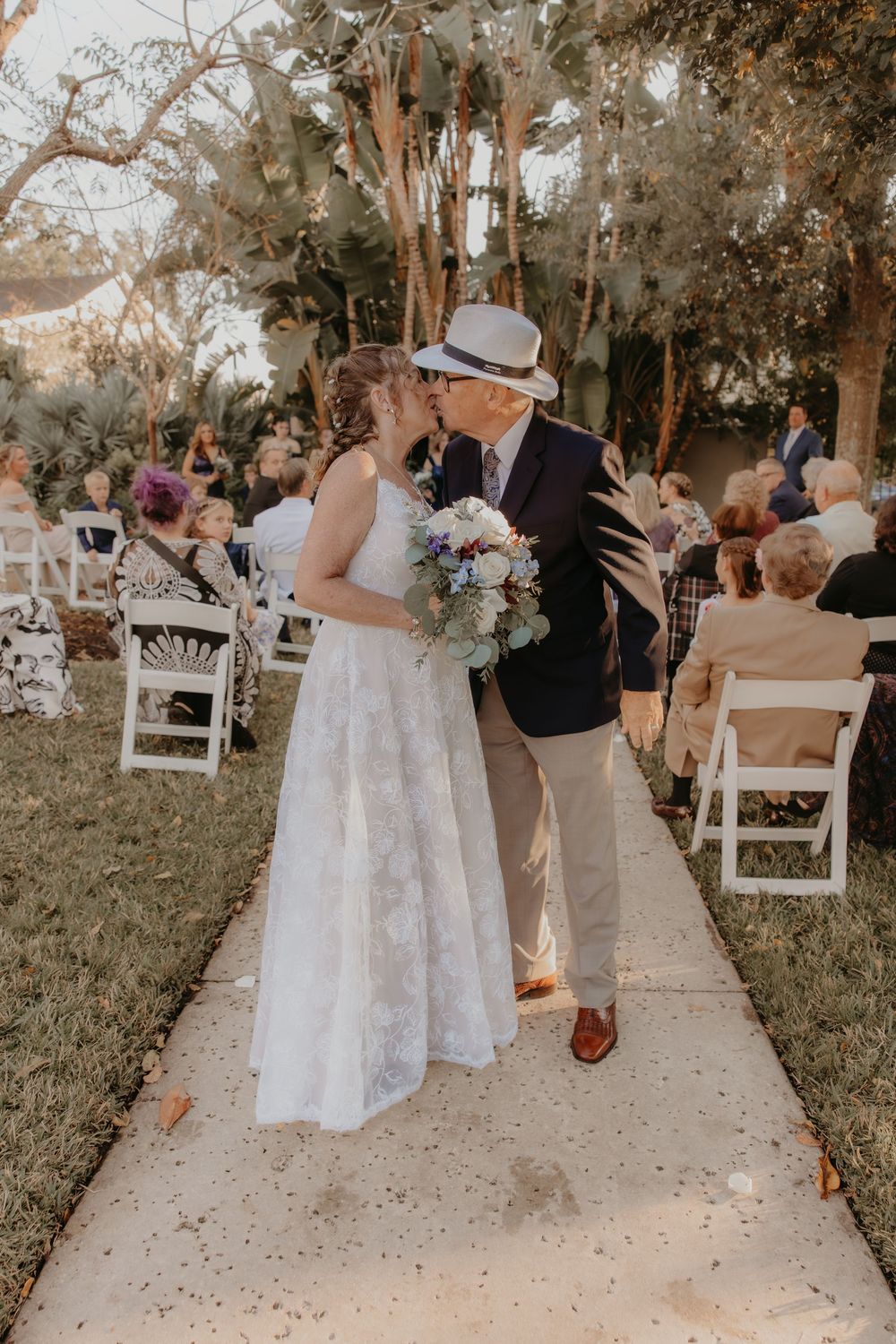 A romantic outdoor wedding ceremony with white chairs and tall trees as newlyweds share a moment on a concrete path.