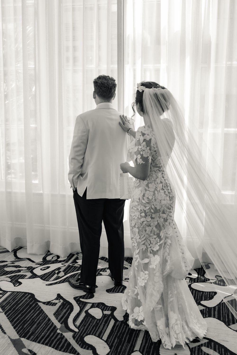 Black and white photo of bride in lace gown with veil and groom in white jacket standing before sheer curtains.