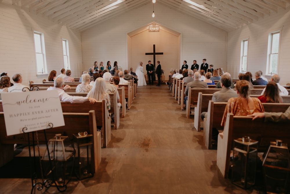 A wedding ceremony inside a rustic white chapel with wooden pews filled with guests facing the altar with a cross.