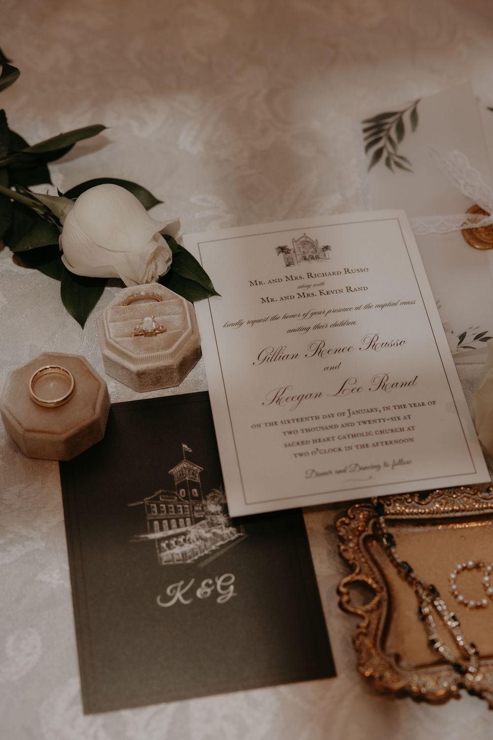 Elegant wedding invitation suite with gold rings, velvet ring boxes, white rose, and monogrammed K&G card on floral backdrop.