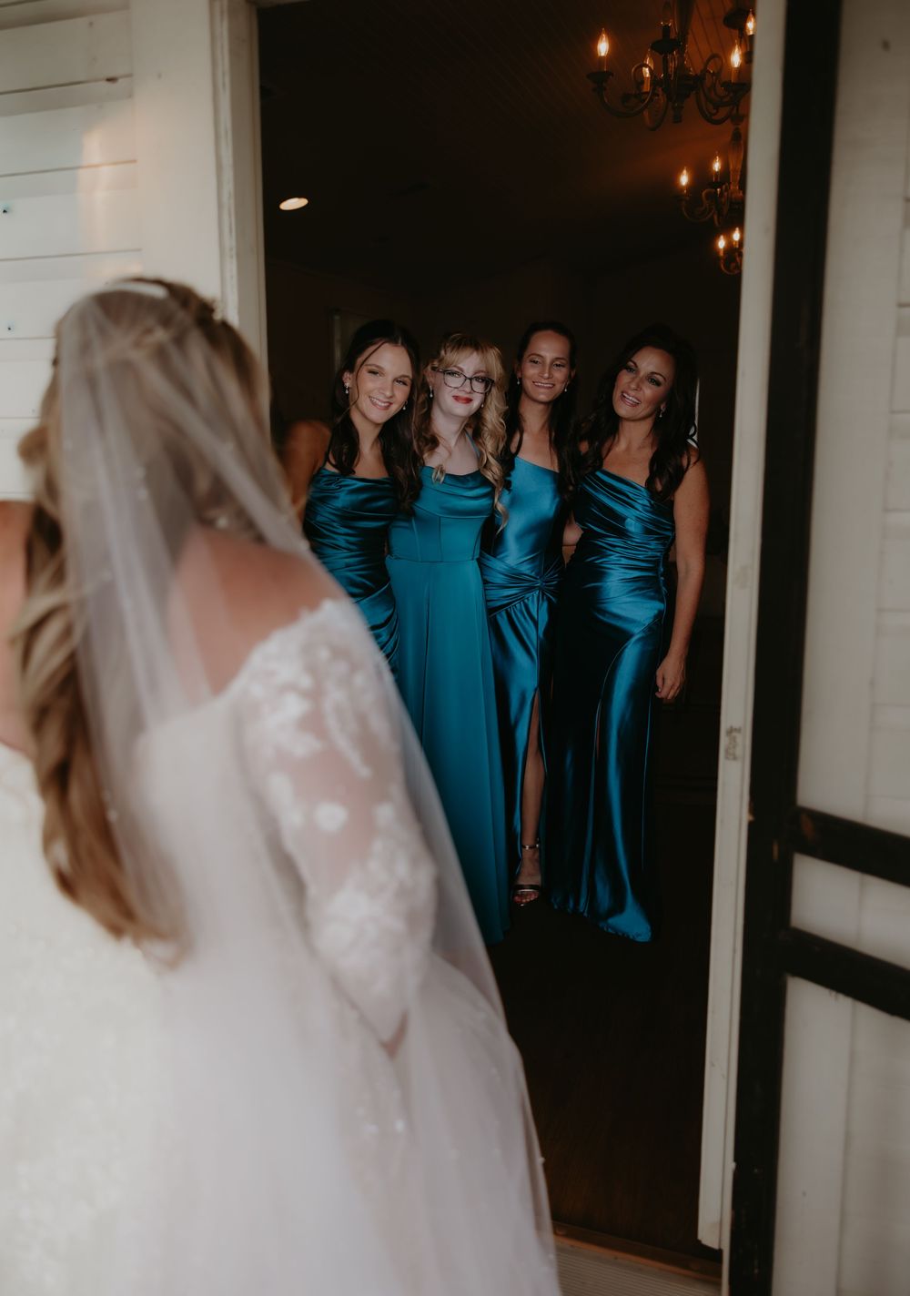 Bride in white lace gown with veil seen from behind as bridesmaids in teal satin dresses react with joy.