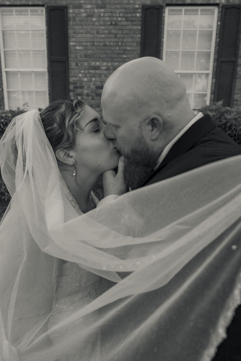 Black and white photo of a bride and groom sharing a romantic kiss, with her veil flowing, outside a brick building.