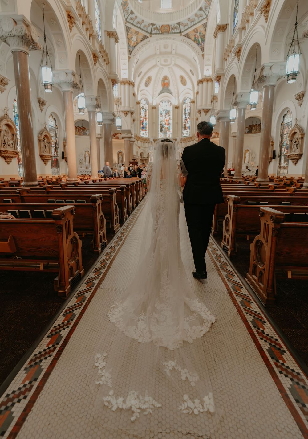 Bride in lace gown with long veil walks down ornate church aisle with escort during wedding ceremony.