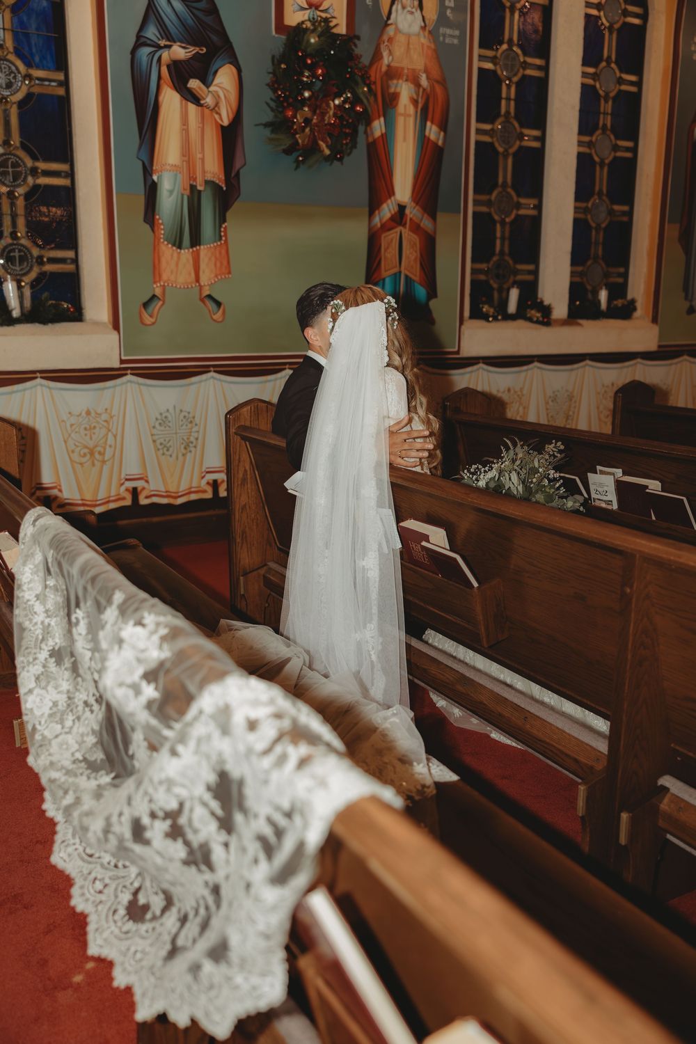Bride and groom share a kiss at the altar inside a church decorated with religious icons and wooden pews.