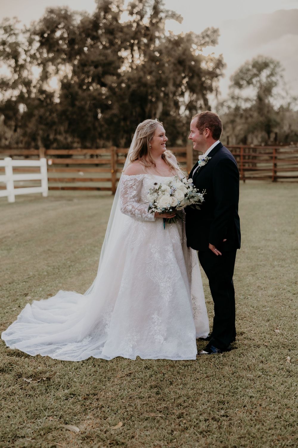 A couple shares intimate moments in their wedding attire on a grassy field with white fencing in the background.