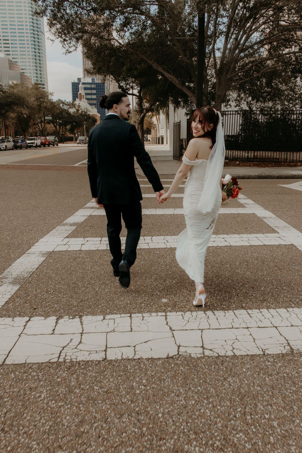 Newlywed couple holding hands walking through a crosswalk in the city, bride in white gown with veil holding bouquet.