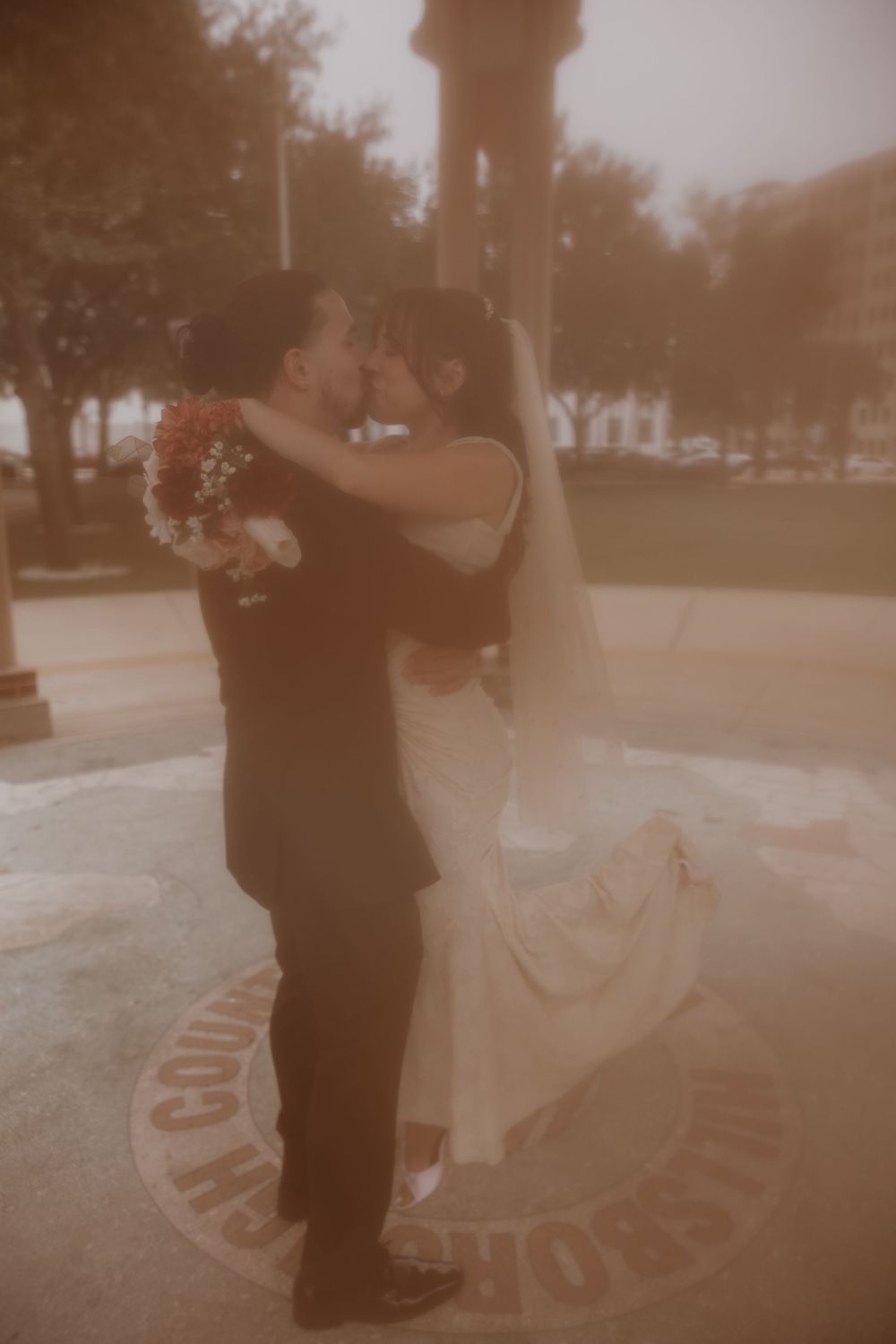 A couple shares a romantic dance moment in their wedding attire on a circular paved area during golden hour.