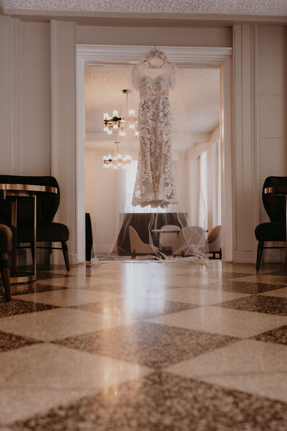 Elegant lace wedding dress hanging in a grand hotel doorway with marble floors and chandelier lighting.