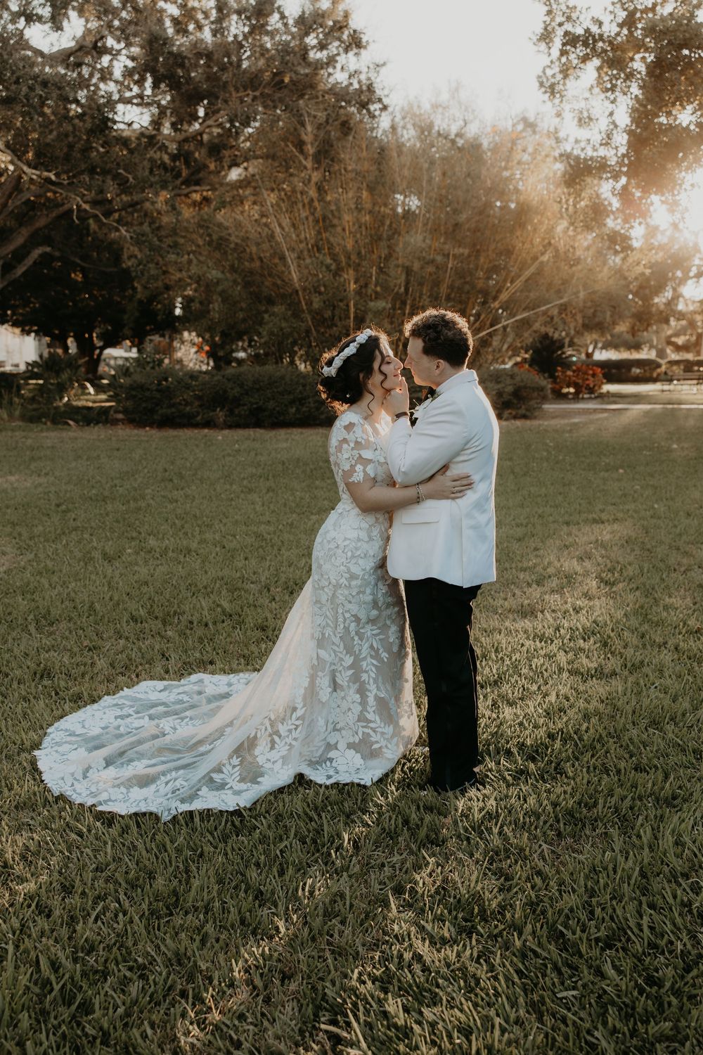 A bride and groom share a romantic kiss at golden hour on a lush green lawn, her lace gown trailing beautifully.
