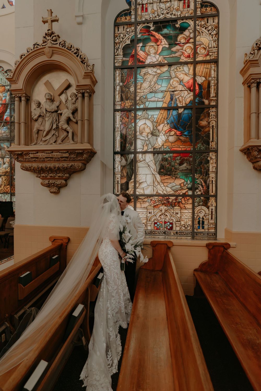 Bride and groom stand before a stunning stained glass window in a historic church, with wooden pews and ornate religious sculptures.