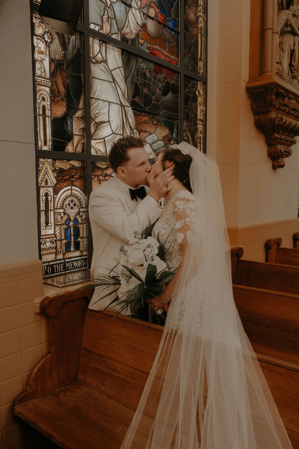 Bride and groom share a kiss in a church pew with a stunning stained glass window backdrop on their wedding day.