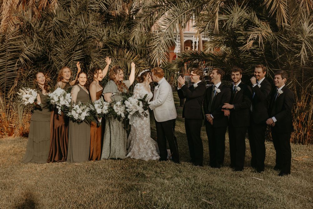 Wedding party poses outdoors under palm trees, bridesmaids in earthy tones holding white bouquets beside groomsmen in black suits.