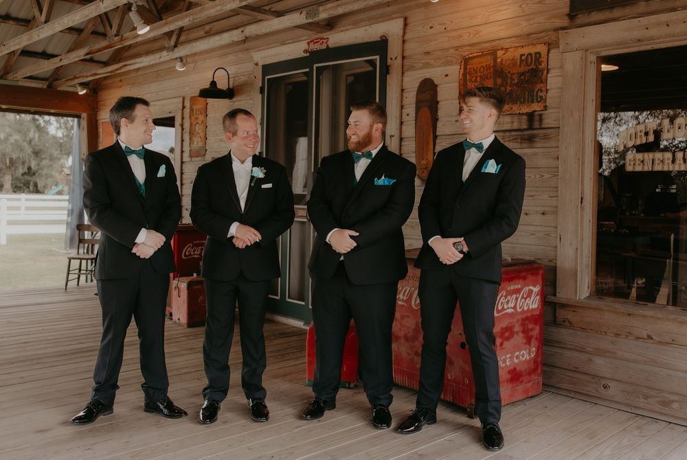 Four groomsmen in black tuxedos with teal bow ties stand near vintage Coca-Cola crates at a rustic wedding venue.