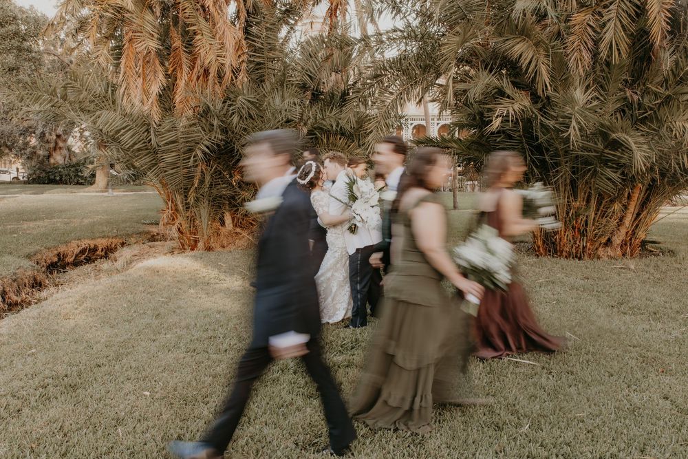 Blurred motion shot of wedding party walking across green lawn with tropical palm trees in background.
