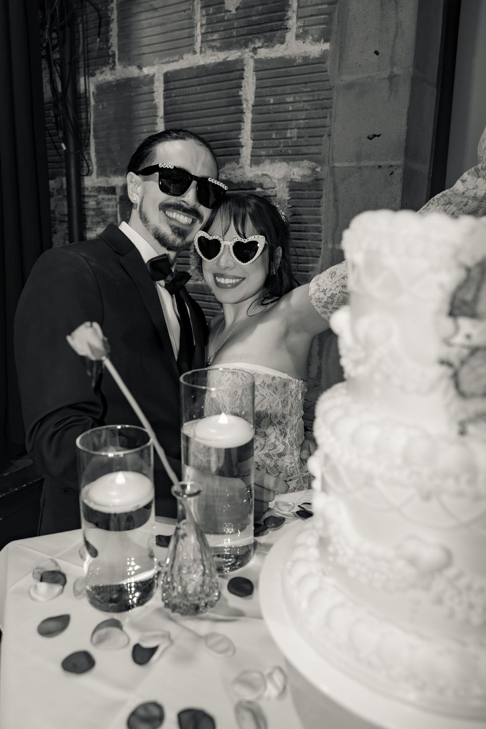 Newlyweds wearing fun sunglasses smile beside their wedding cake in black and white photo.
