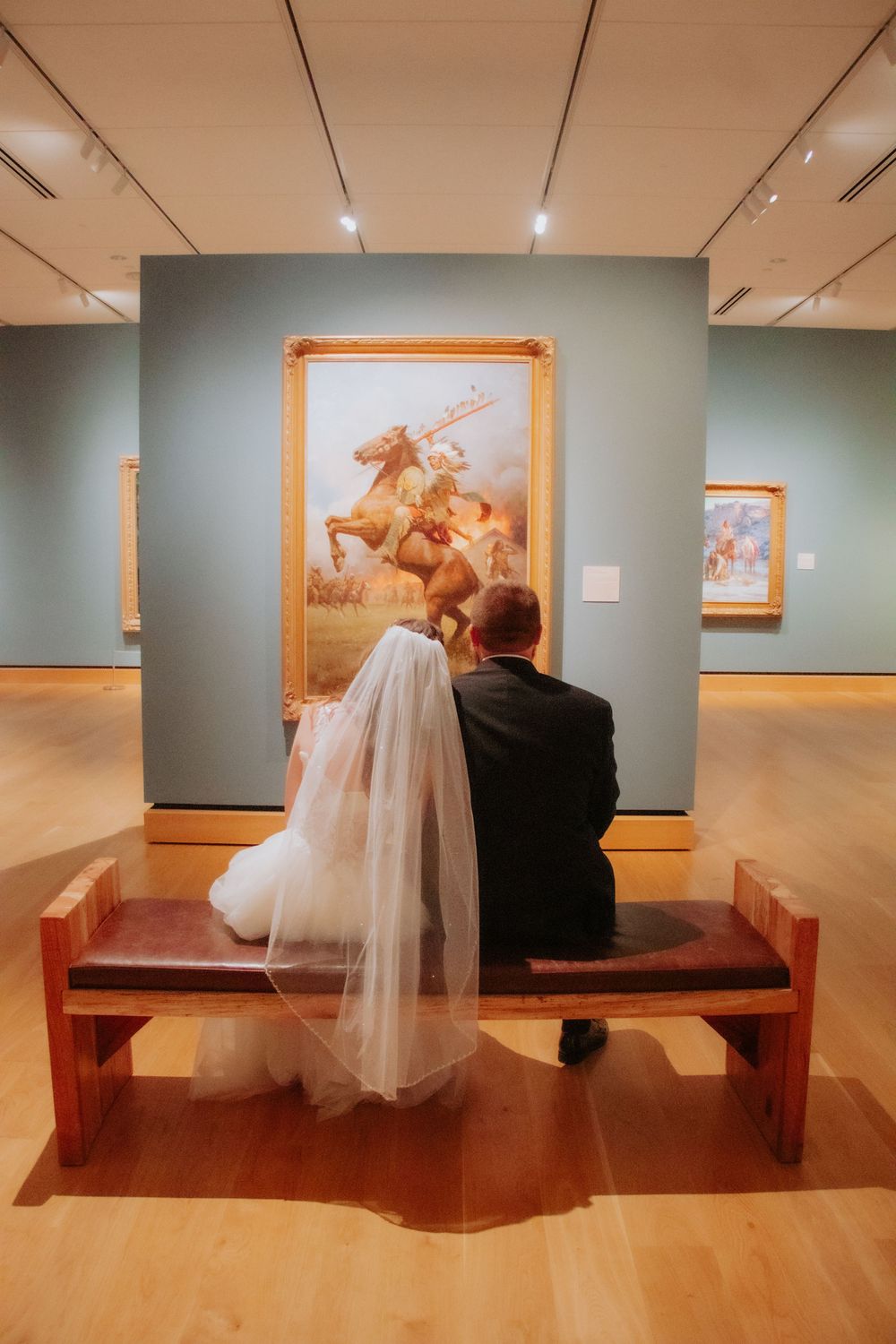Newlywed couple in wedding attire sitting on a museum bench, admiring a large classical painting on a teal wall.