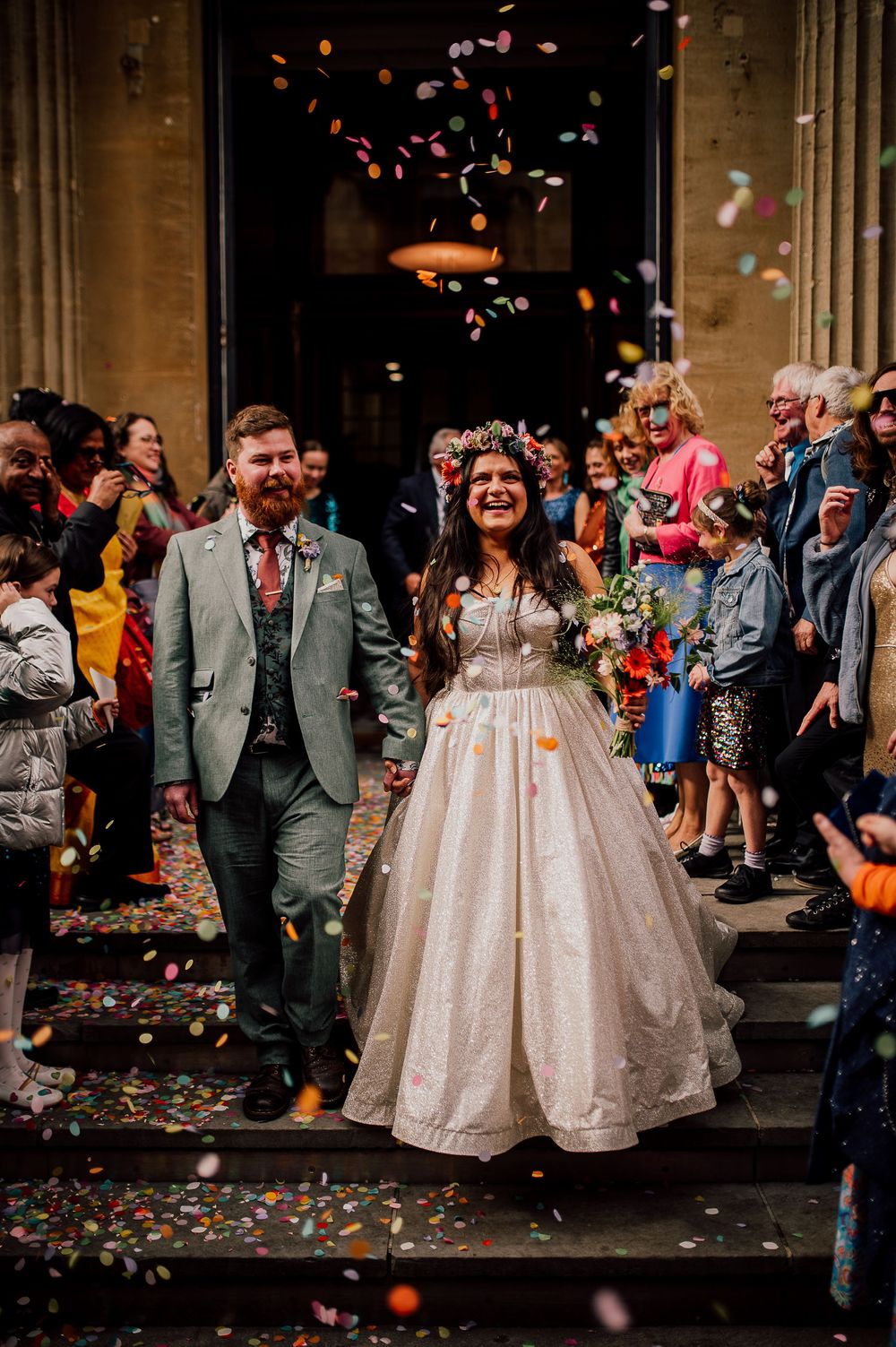 Wedding guests throw colorful confetti as newlyweds exit church in elegant champagne gowns and formal attire.