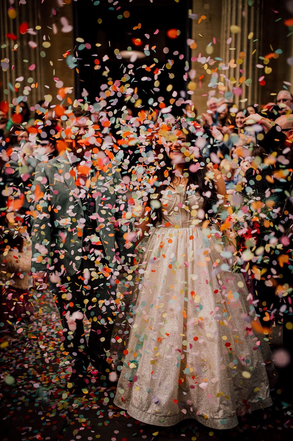 A white lace wedding dress photographed in a sequence with colorful confetti falling around it in dramatic lighting.