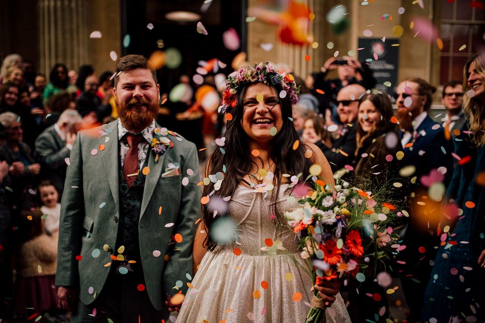 Joyful couple laughs while being showered with vibrant confetti outside a historic building.