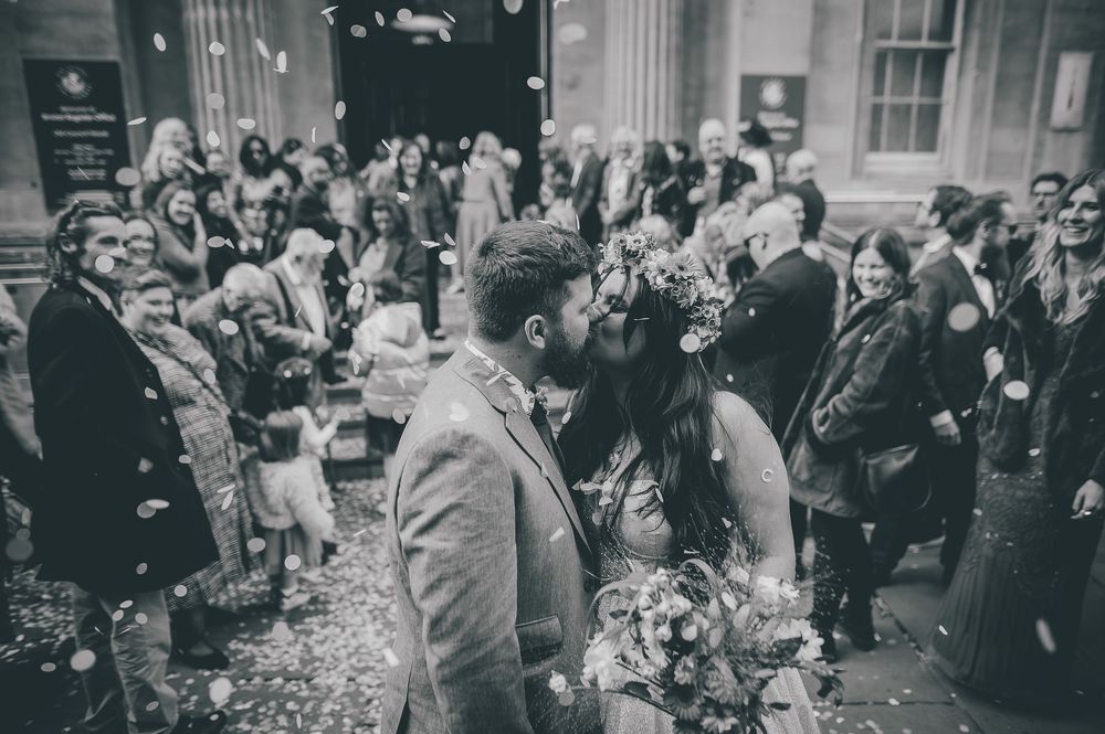 A joyful crowd celebrates with confetti and cheers outside a building during what appears to be a wedding celebration.