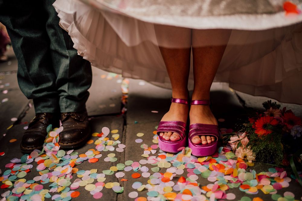 Pink sandals and dark shoes stand on confetti-covered floor at a celebration event.
