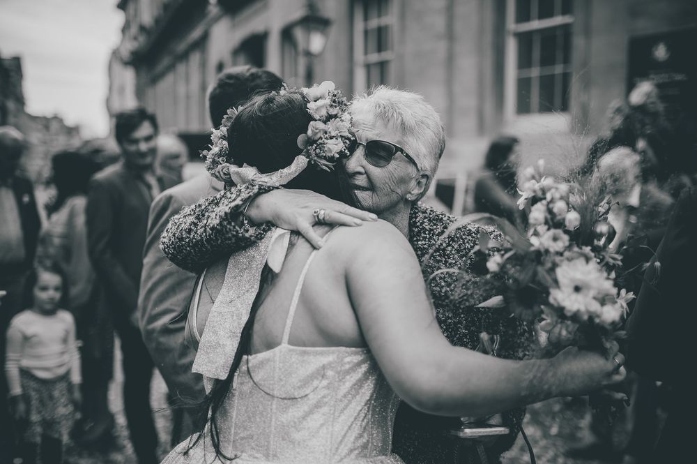 A vintage black and white photograph shows an emotional embrace at a wedding celebration with flowers and onlookers.