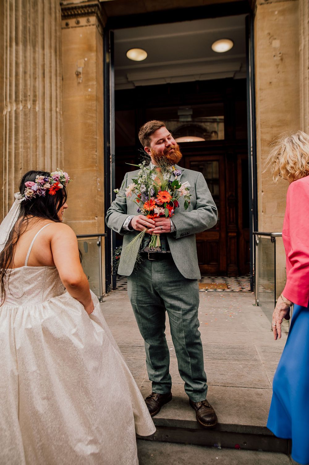 A couple shares a joyful moment outside a historic building entrance as the groom holds a vibrant floral bouquet.
