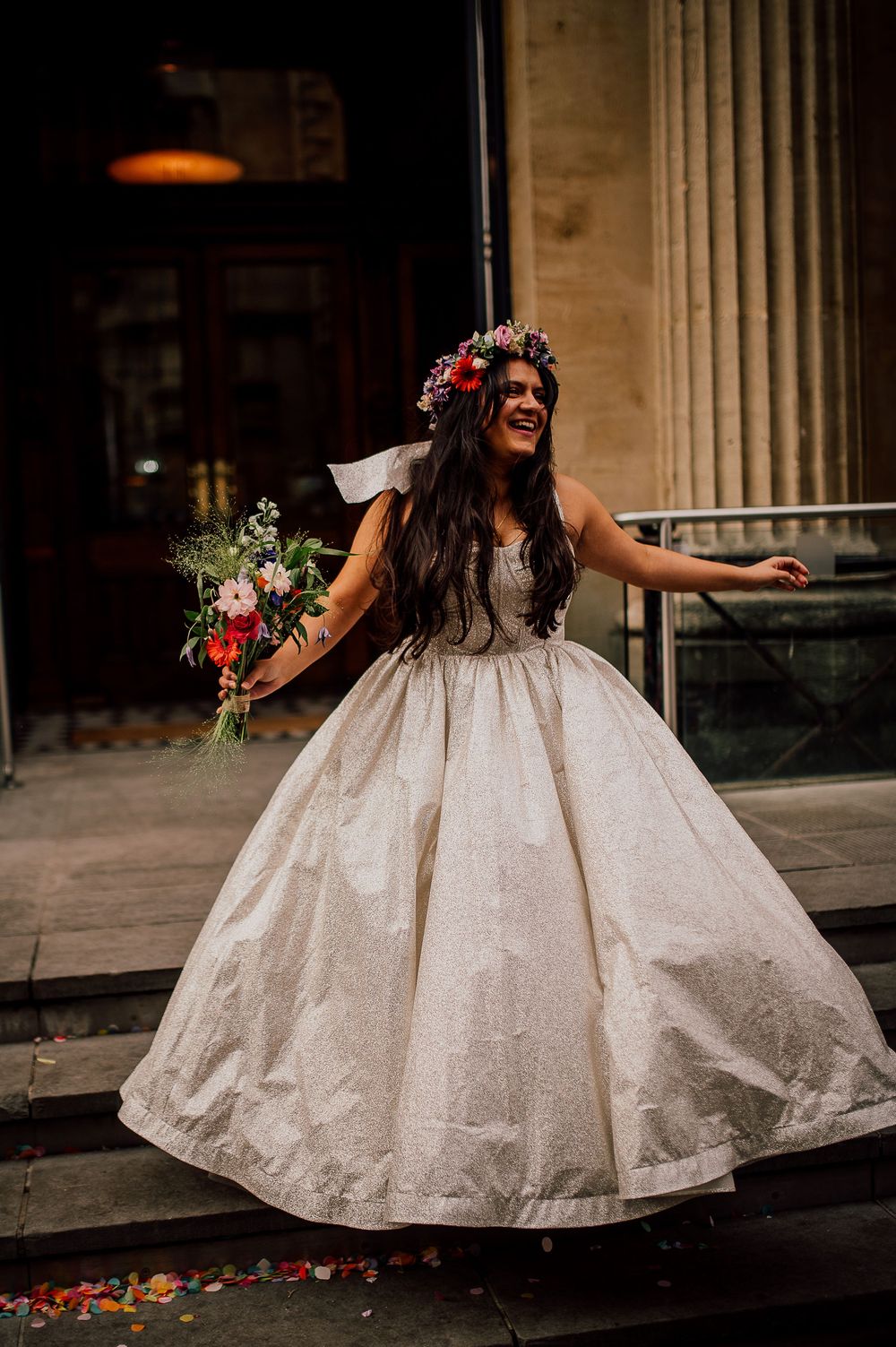 A bride in a flowing white ballgown and floral crown twirls joyfully on the steps of a grand building.