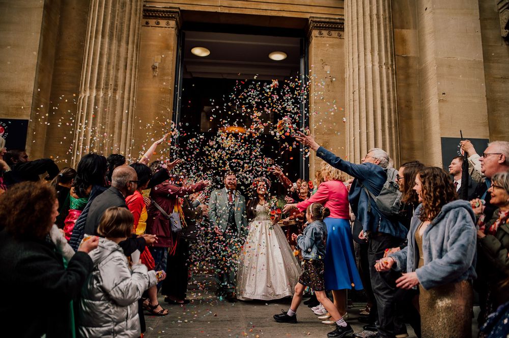 Wedding guests shower newlyweds with colorful confetti as they exit a grand columned building entrance.