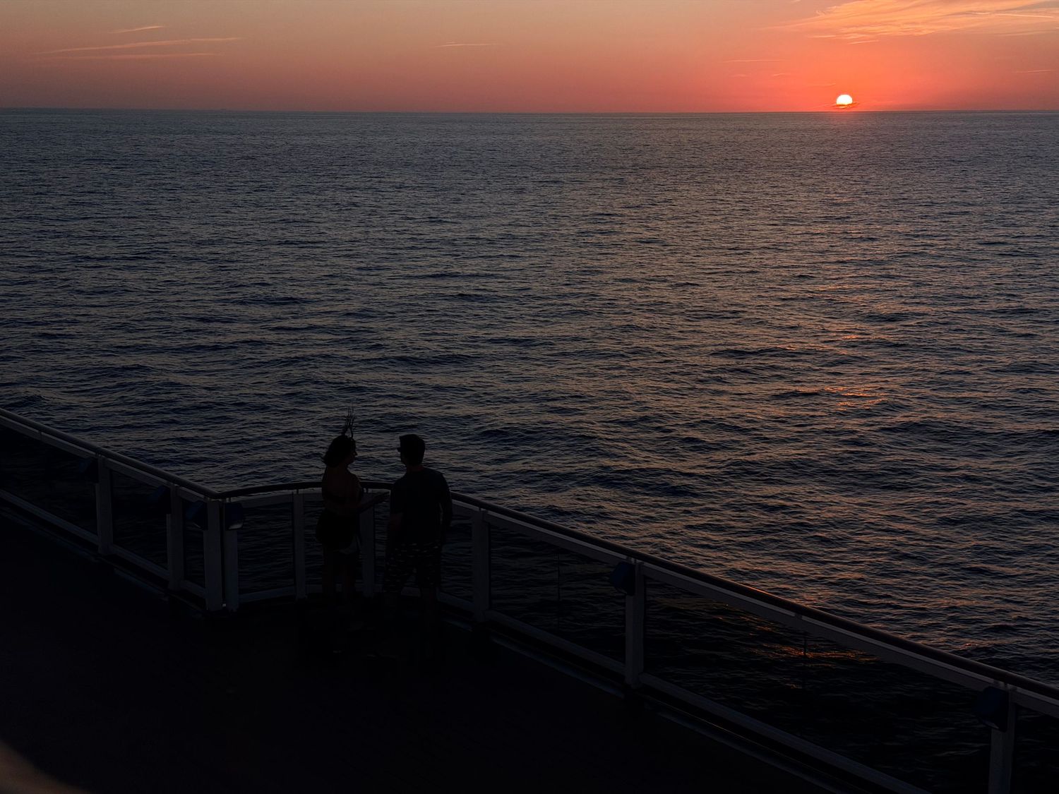Two people watch a vibrant orange sunset over calm ocean waters from a cruise ship deck railing.