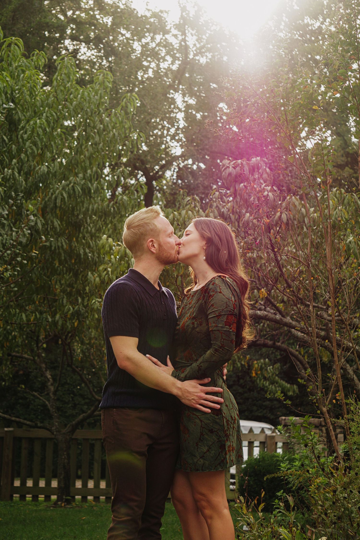 A couple embraces and kisses in a garden setting with flowering trees and sunlight filtering through the foliage.