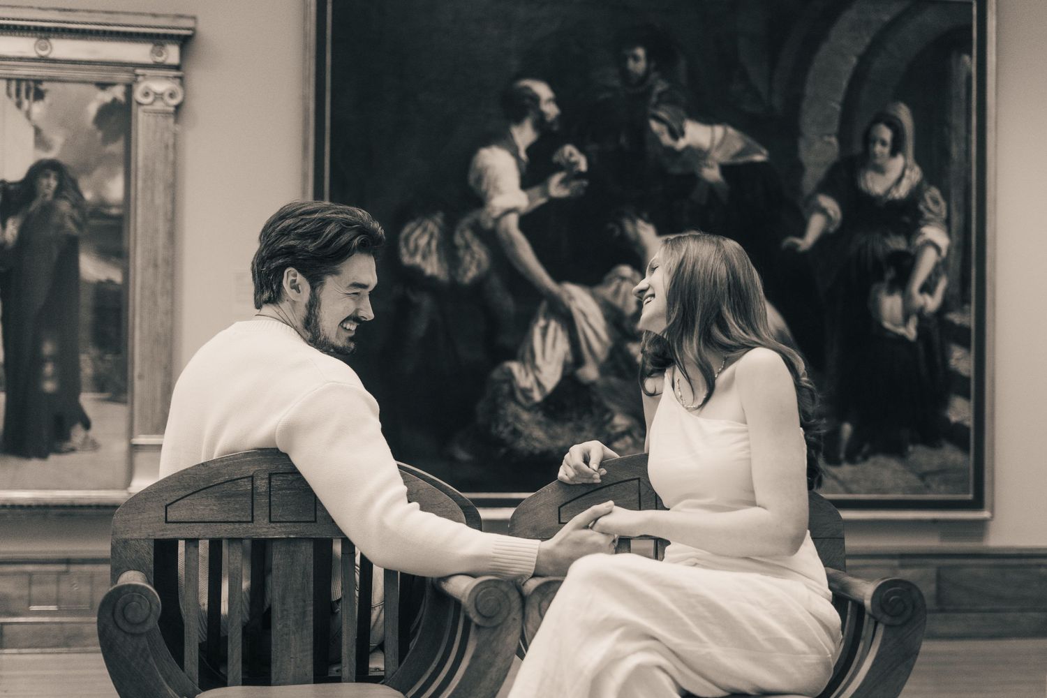 Couple holding hands in a museum, seated before a large classical painting in a romantic black and white photo.