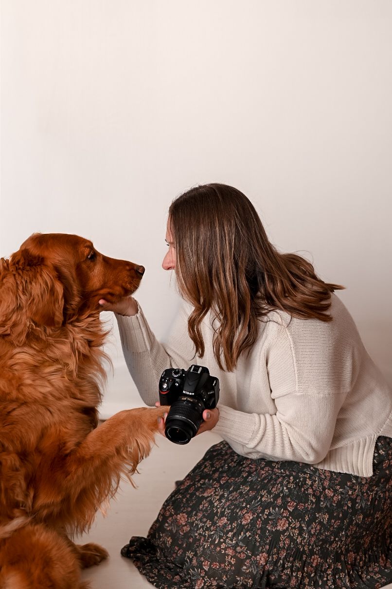 Woman in white sweater holds Nikon camera while sharing a tender moment with a golden Irish Setter dog.