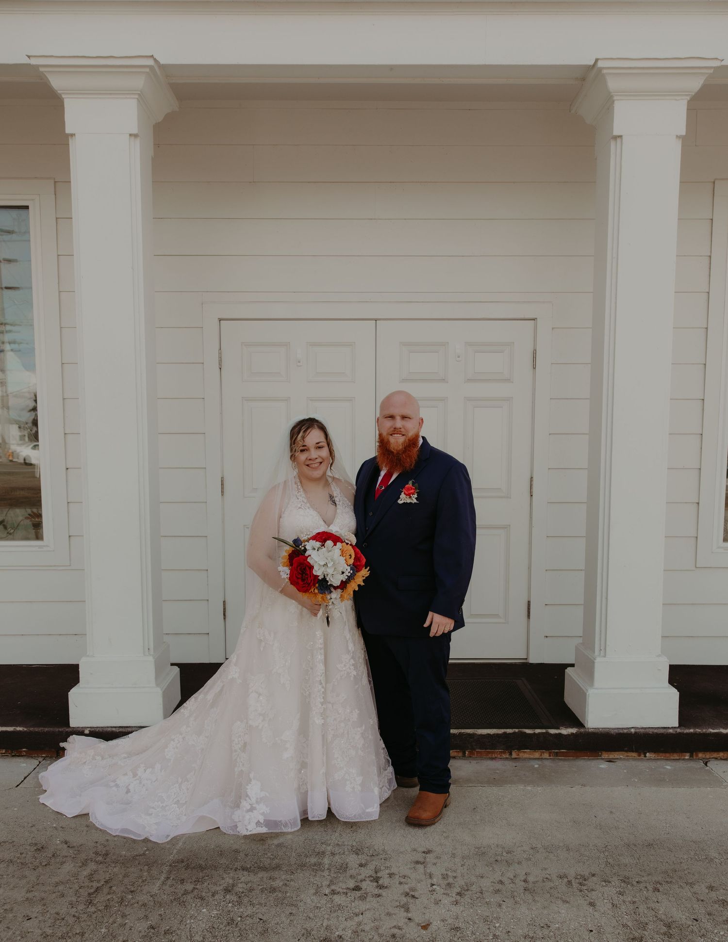Bride in lace gown holding colorful bouquet stands with groom in navy suit and red beard before white chapel doors.