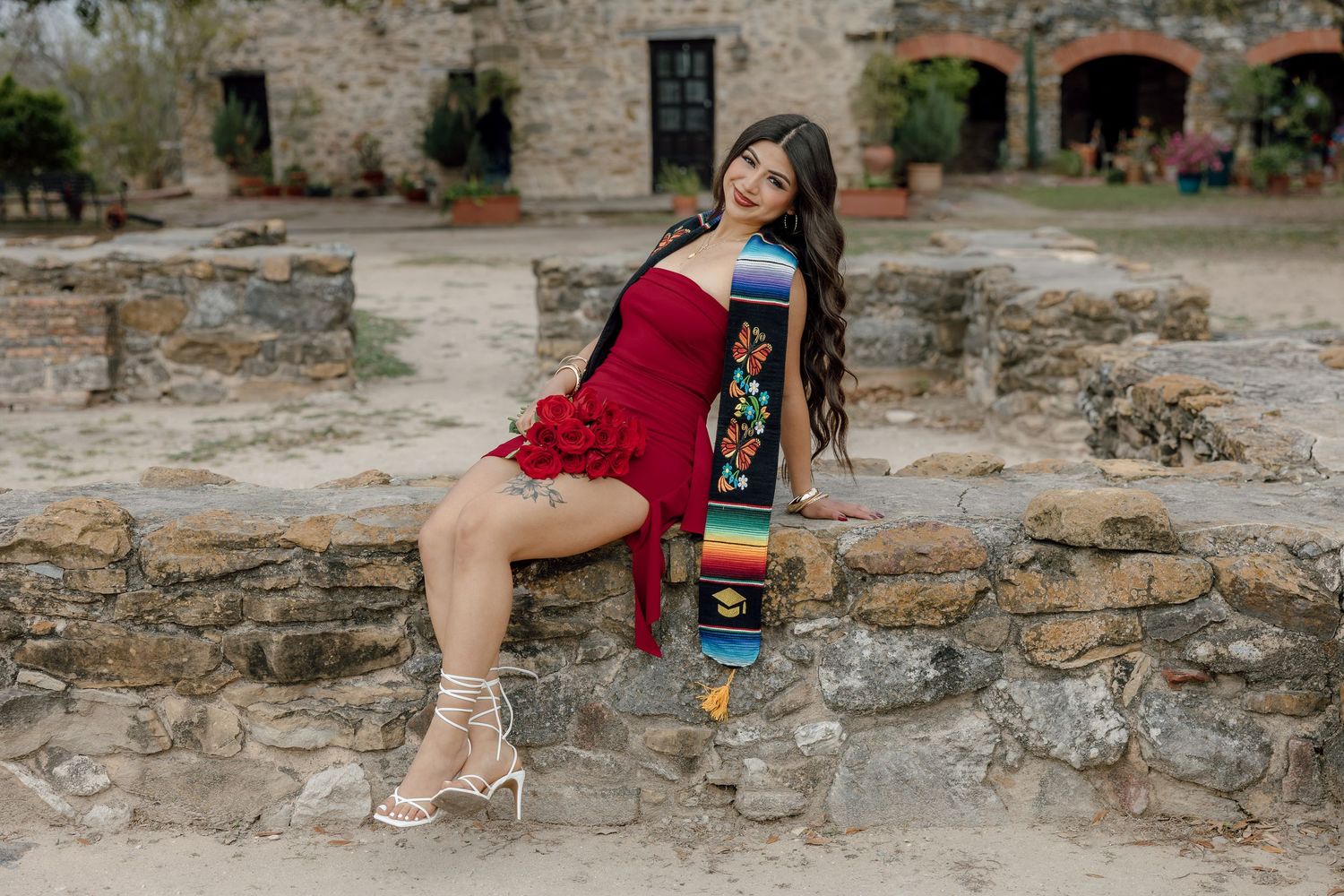 Texas senior photo of girl in red dress sitting on stone wall with graduation stol