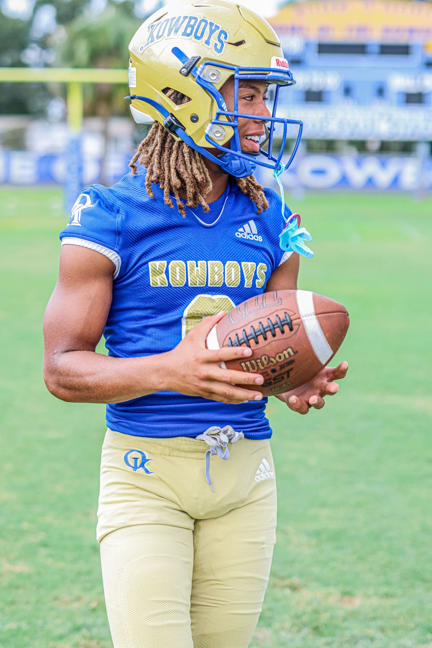 Football player in blue and gold Cowboys uniform holds ball during practice on grass field.