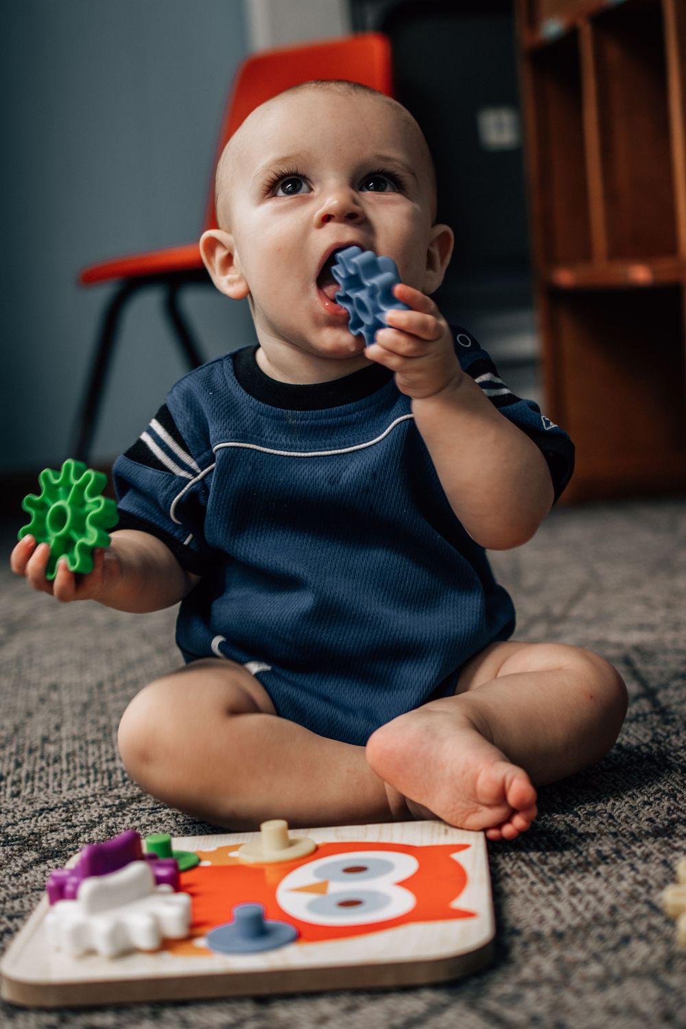 An adorable baby in a navy onesie sits on a carpet chewing a blue toy gear with puzzle pieces nearby.
