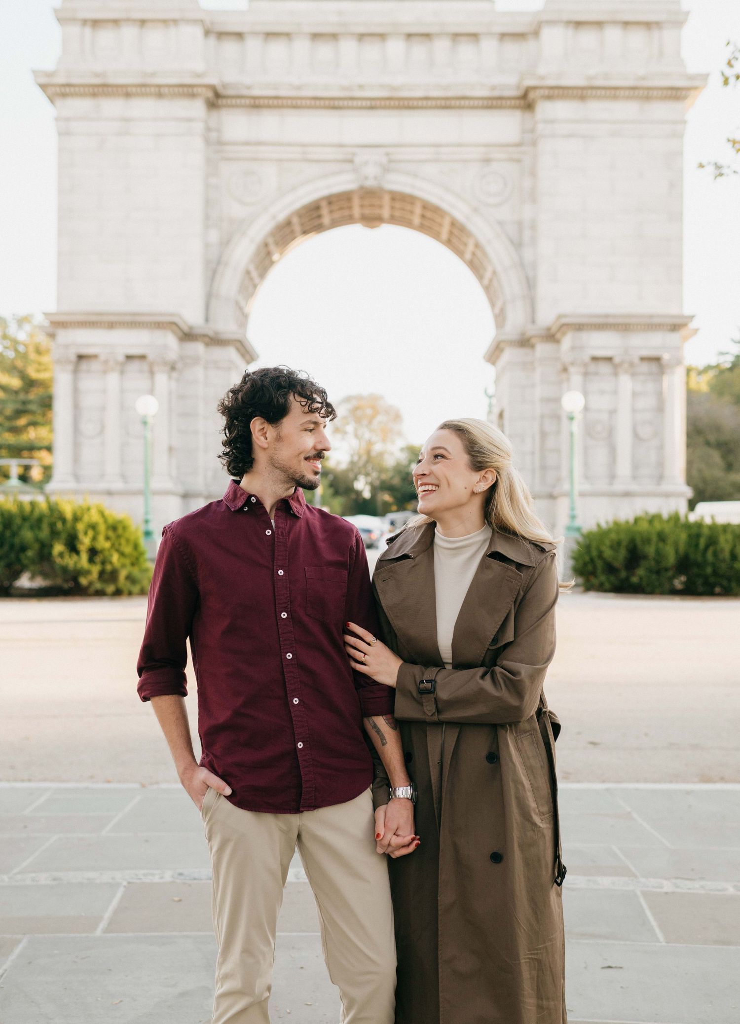 A happy couple shares a romantic moment while walking together near a grand stone archway on a sunny day.