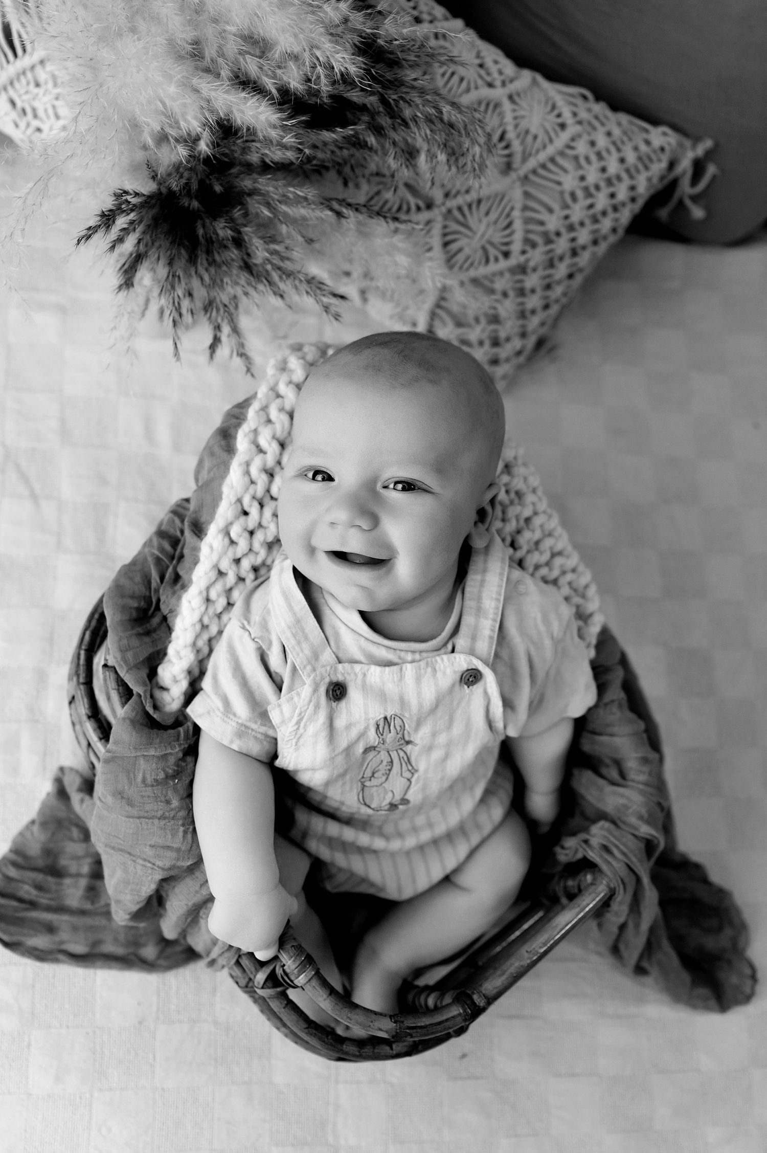 Baby wearing blue denim dress with ruffled collar sits on white blanket smiling at camera.