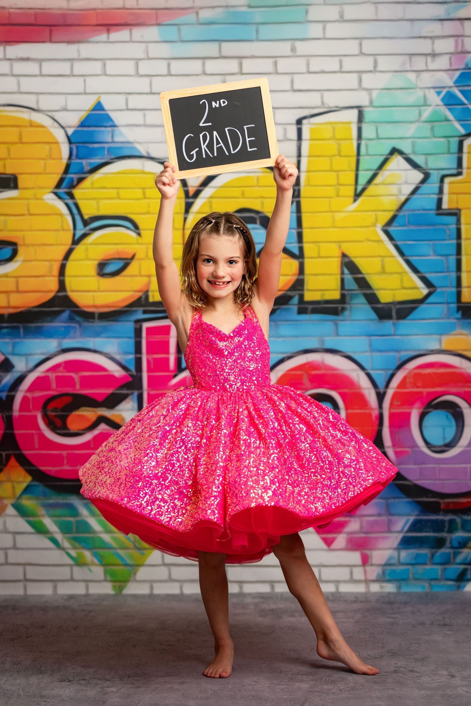 A bright pink tulle dress twirls against a colorful graffiti wall with a chalkboard sign showing second grade.
