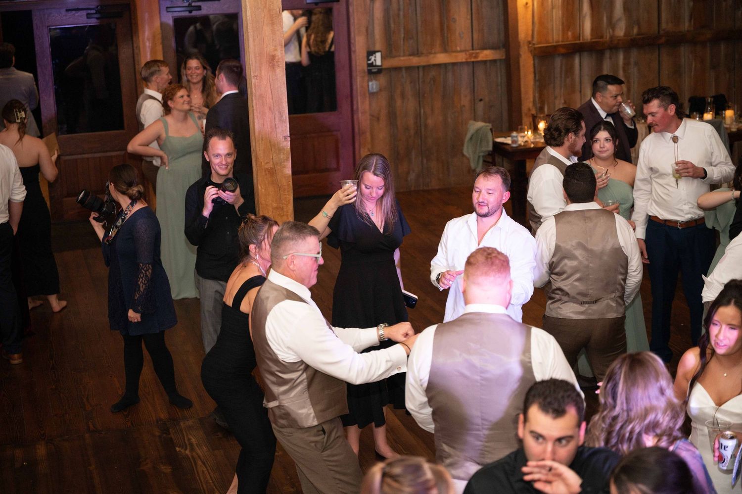 Wedding guests dance and celebrate on a wooden dance floor at an evening reception with warm lighting.